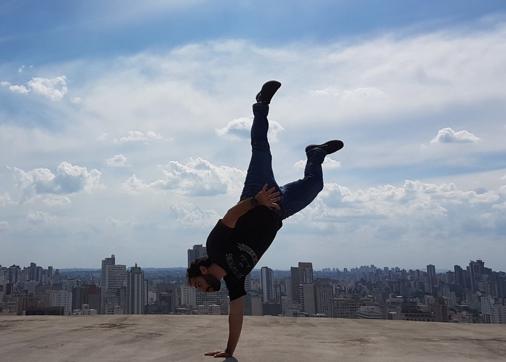 É uma imagem do autor do blog plantando bananeira com uma mão só na laje do prédio do shopping italia em Curitiba. O céu está azul com algumas nuvens brancas ao fundo.