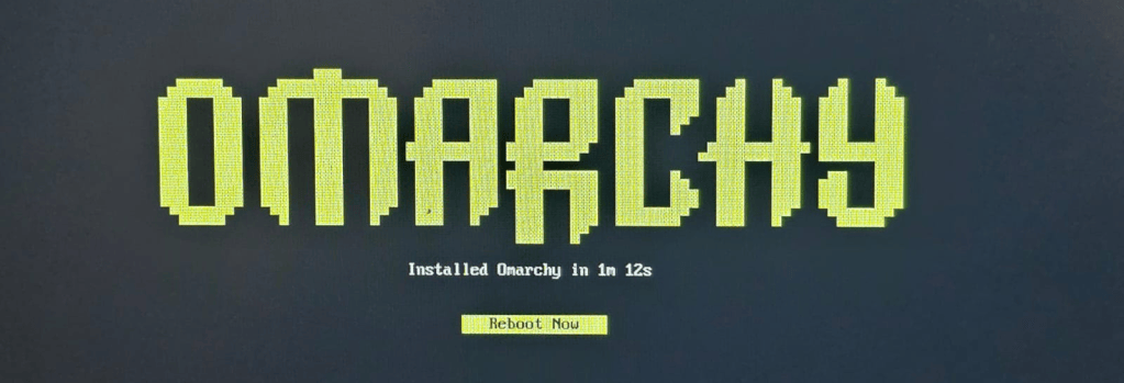 Tela final do instalador do Omarchy, com o logo em letras verdes pixeladas, a mensagem "Installed Omarchy in 1m 12s" e o botão Reboot Now